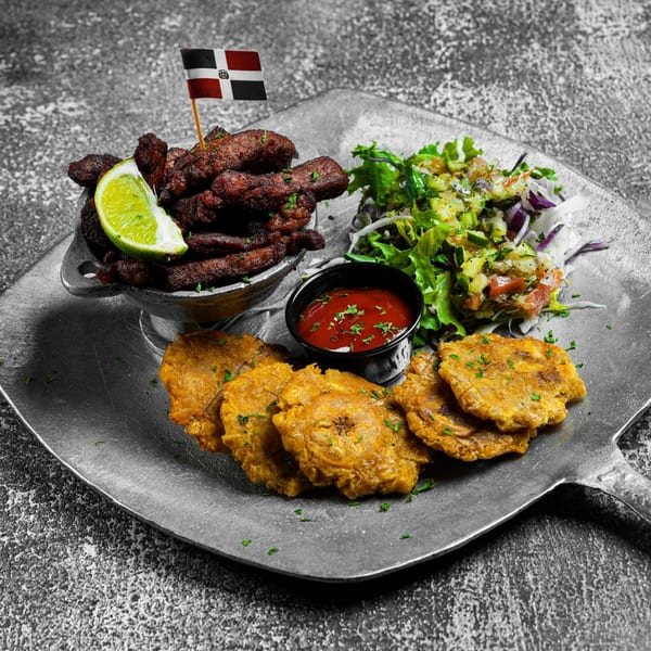 Dominican fried pork, tostones, and salad with a lime wedge and flag on a metal plate.