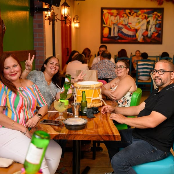 Four happy diners at Perico Ripiao Latin Food, with drinks, a drum, and a colorful painting.