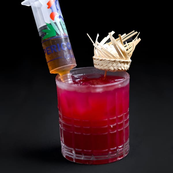 A vibrant red cocktail with a Perico Ripiao branded syringe and a straw hat garnish.