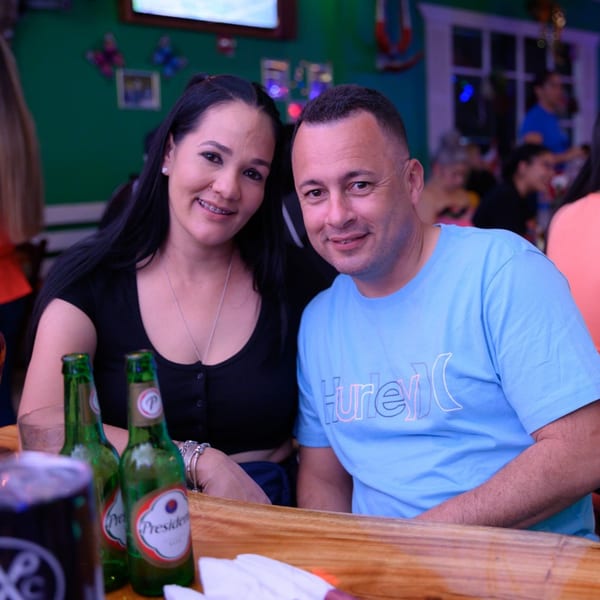 A couple smiles at the camera, with two Presidente beer bottles on a wooden table.