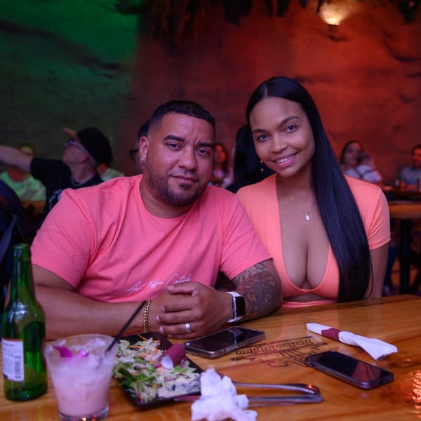 A couple smiles at the camera, seated at a table with drinks and salad in a dimly lit restaurant.