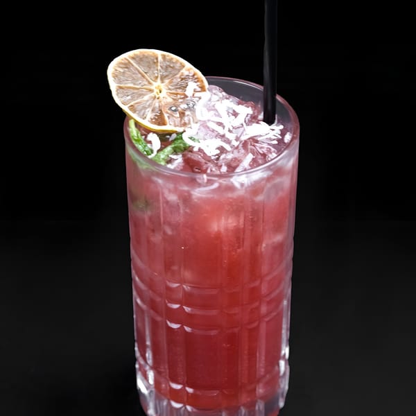 A vibrant red cocktail in a textured glass, garnished with a dried lemon slice and shredded coconut.