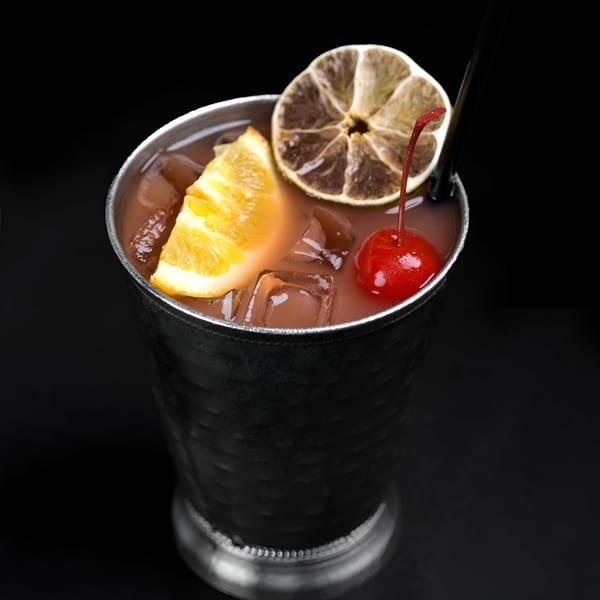 A Perico Ripiao cocktail in a metal cup, garnished with orange, dried lime, and a cherry.